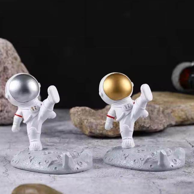 Two astronaut phone stand models with silver and gold helmets on textured moon surface bases