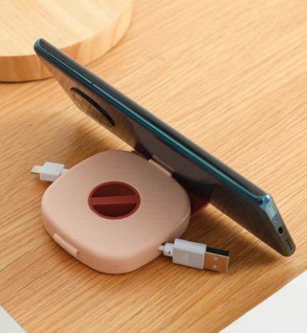 Compact USB cable holder in beige with a phone stand feature holding a smartphone on a wooden surface