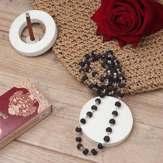 Ebony wood mala beads with silver accents displayed on a white holder beside a red rose and incense stick