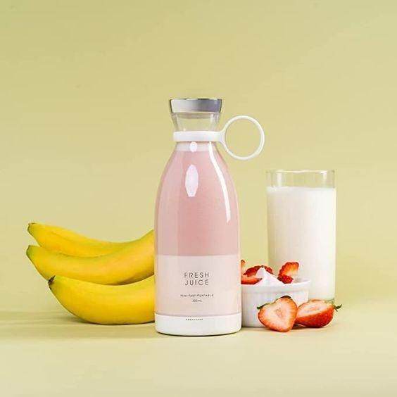 Portable mini juice blender with pink smoothie next to bananas, strawberries, and a glass of milk
