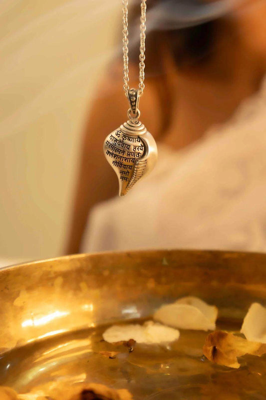 Silver Shri Krishna conch pendant necklace with Sanskrit inscriptions hanging above brass bowl with flower petals